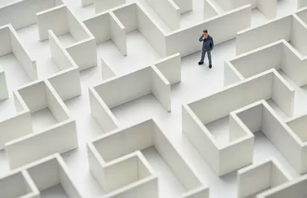 A businessman stands in a large white maze, contemplating his path.