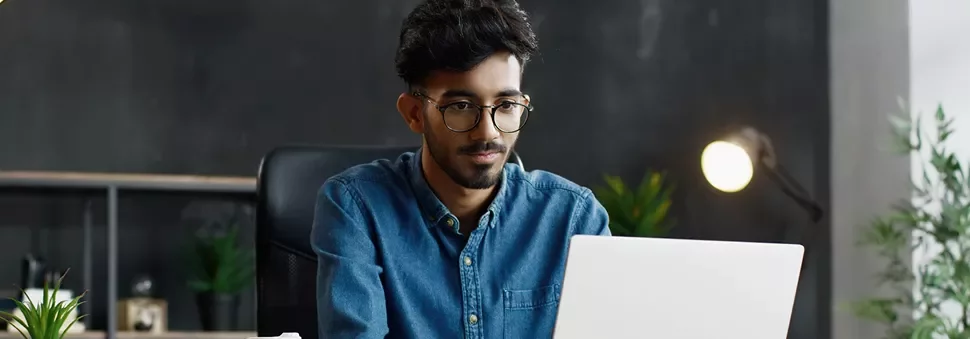 Man in glasses working on a laptop at a desk with plants and a lamp.