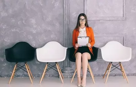 Woman in orange blazer sitting in a chair holding a clipboard.