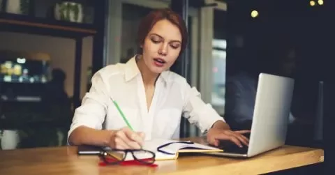 Woman working on a laptop and writing in a notebook.