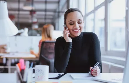 Woman in office using phone and writing notes.