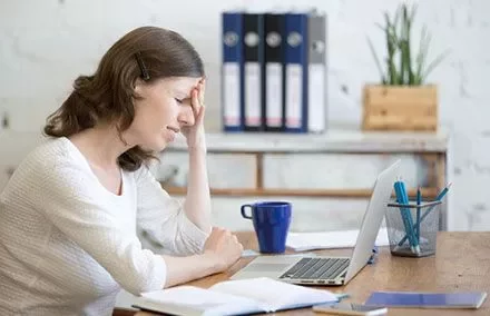 Woman at desk with hand on head, looking stressed at laptop.