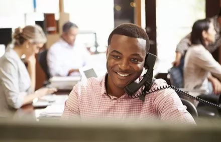 Man on phone smiling at desk in a busy office.