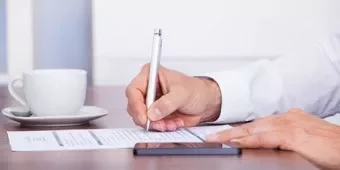 Person writing on a paper beside a cup and smartphone.