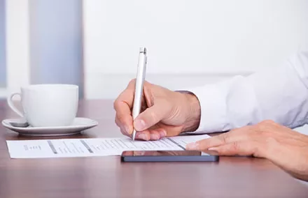 Person signing document at a desk with a cup of coffee and a phone.