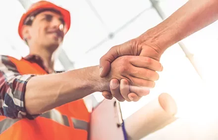 Construction worker shaking hands with a colleague, holding blueprints.