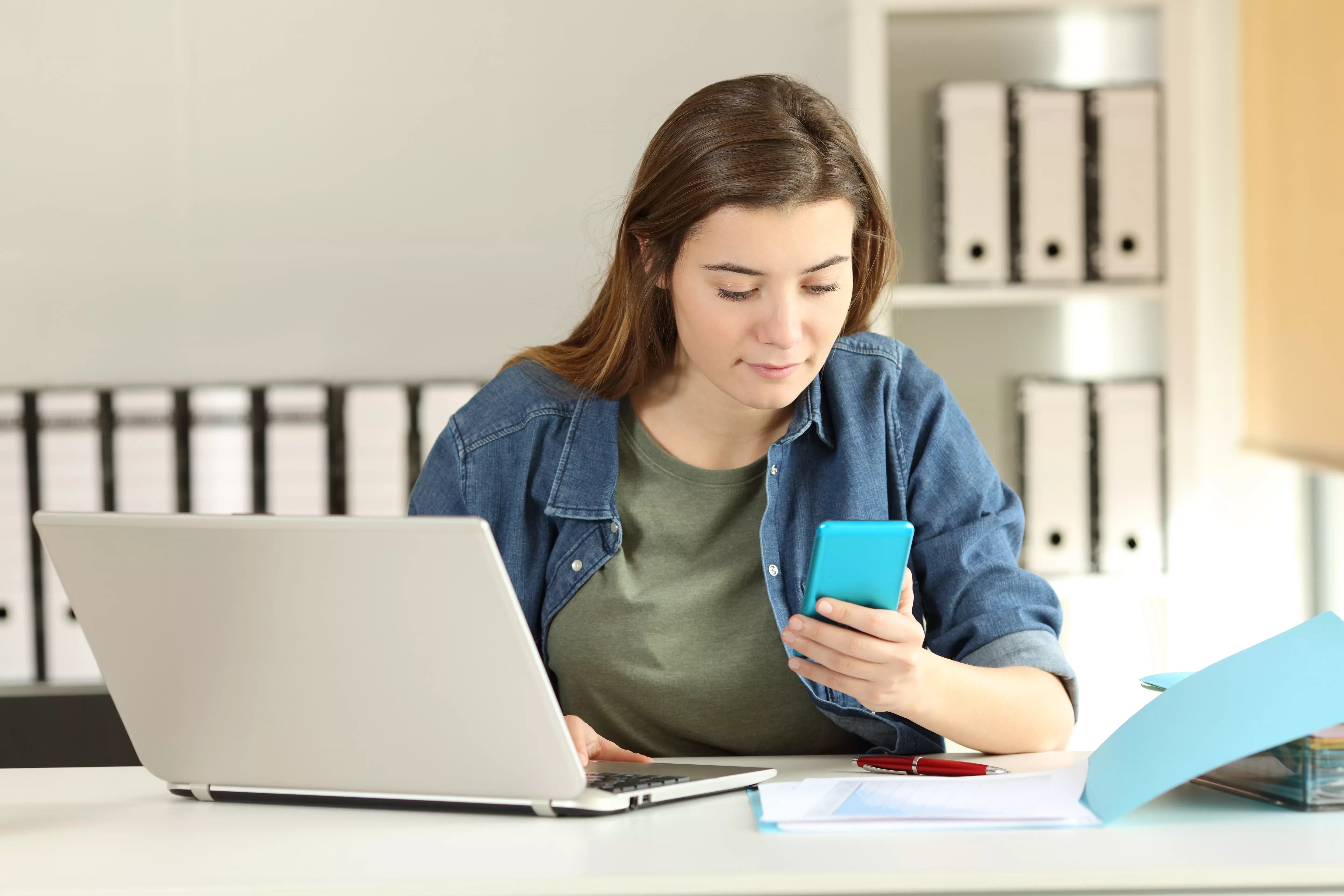 Person using a smartphone while working on a laptop at a desk.