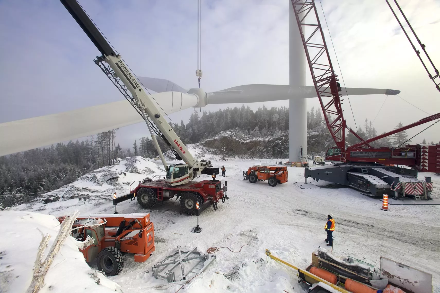 Workers installing a wind turbine blade with cranes in a snowy landscape.
