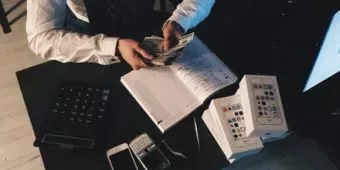 Person counting money, with phones and calculator on desk.
