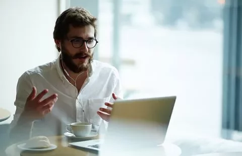 Man with glasses using laptop and headset in cafe.